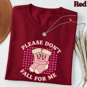 Puede incluir: Una camiseta burdeos de cuello redondo con el texto "PLEASE DON'T FALL FOR ME" y un gráfico de dos calcetines abrazándose. Un collar dorado con un colgante está drapeado sobre la camisa. La palabra "Red" está en la esquina superior derecha.