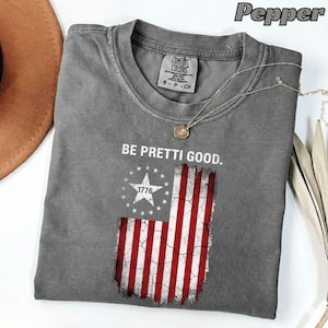 May include: A gray t-shirt with the text "BE PRETTI GOOD." and a distressed American flag graphic. The flag design features a white star with "1776" and red and white stripes. A silver necklace with a pendant is on the shirt.