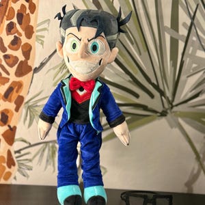 May include: A plush doll of a character with a mischievous expression, wearing a blue suit with a red bow tie and black shirt. The doll has black hair and green and blue eyes. Black-framed glasses are next to the doll. The doll is approximately 10 inches tall.