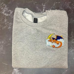 May include: A gray sweatshirt with a crew neck. The sweatshirt features an embroidered design of a cartoon dragon on a cloud with a crescent moon and stars. The dragon is orange, blue, and yellow.