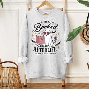May include: A light gray crewneck sweatshirt featuring a graphic of a white ghost wearing heart-shaped sunglasses, reading a book titled PLANS, and holding a glass of wine. The text reads SORRY, I'M Booked FOR THE AFTERLIFE OCTOBER SCHEDULE IS FULL.