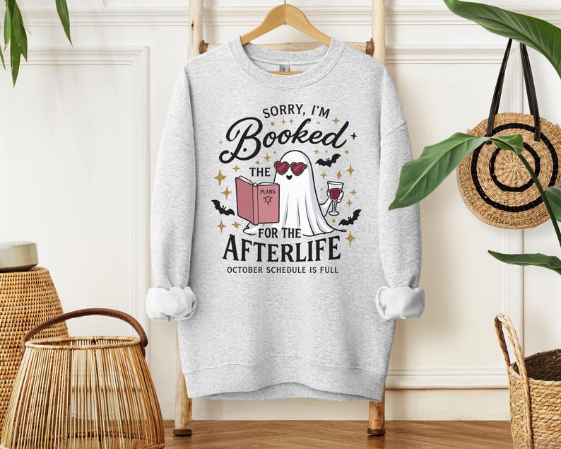 May include: A light gray crewneck sweatshirt featuring a graphic of a white ghost wearing heart-shaped sunglasses, reading a book titled PLANS, and holding a glass of wine. The text reads SORRY, I'M Booked FOR THE AFTERLIFE OCTOBER SCHEDULE IS FULL.