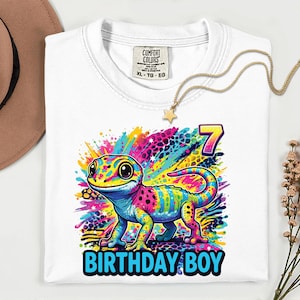 White t-shirt with a colorful gecko graphic. The gecko is depicted with vibrant hues of pink, blue, yellow, and green. The number 7 and the words "BIRTHDAY BOY" are also featured on the shirt.