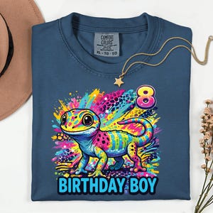 May include: A blue t-shirt with a colorful gecko graphic and the text "BIRTHDAY BOY" in blue. The gecko is multi-colored with pink, yellow, blue, and green. The number "8" is above the gecko. A gold necklace with a star pendant is on the shirt.