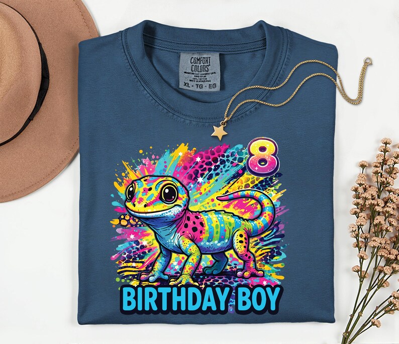 May include: A blue t-shirt with a colorful gecko graphic and the text "BIRTHDAY BOY" in blue. The gecko is multi-colored with pink, yellow, blue, and green. The number "8" is above the gecko. A gold necklace with a star pendant is on the shirt.