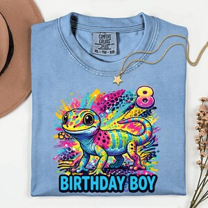 May include: A light blue t-shirt with a colorful gecko graphic and the text "BIRTHDAY BOY" in blue. The gecko is multi-colored with pink, yellow, blue, and green. A gold necklace with a star pendant is on the shirt.