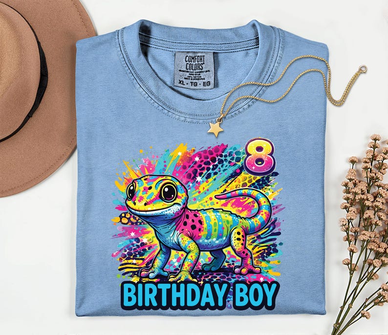 May include: A light blue t-shirt with a colorful gecko graphic and the text "BIRTHDAY BOY" in blue. The gecko is multi-colored with pink, yellow, blue, and green. A gold necklace with a star pendant is on the shirt.
