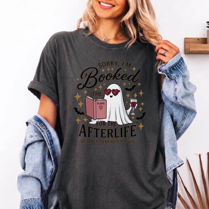 May include: A charcoal gray t-shirt featuring a graphic of a white ghost wearing heart-shaped sunglasses, holding a pink book titled PLANS and a glass of red wine. The text reads SORRY, I'M BOOKED FOR THE AFTERLIFE and OCTOBER SCHEDULE IS FULL.