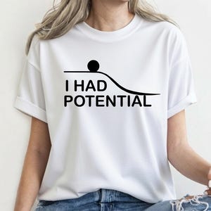 Puede incluir: Una camiseta blanca de manga corta con un gráfico negro de una bola sobre una superficie plana que se curva hacia abajo. Debajo del gráfico, el texto dice I HAD POTENTIAL en una fuente negra, negrita y sin remates.