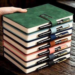 May include: A stack of colorful notebooks with matching pen holders and pens. The notebooks are in shades of green, blue, pink, brown, and black. Each notebook has a pen secured by a matching leather strap and a metal clasp.