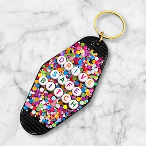 May include: Black motel-style keychain with a gold ring. The keychain is covered in colorful glitter and features the text "DRIVE SAFE BITCH" spelled out with white letter beads. A fun and unique accessory.