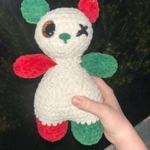 May include: A handmade crochet panda bear with red and green accents. The bear has a white body with glittery thread, red feet and arms, and green ears. It has a black stitched eye and a winking eye.