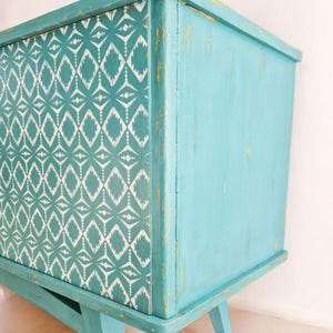 May include: A teal blue cabinet with a geometric pattern on the front. The cabinet has a distressed finish and sits on four legs.