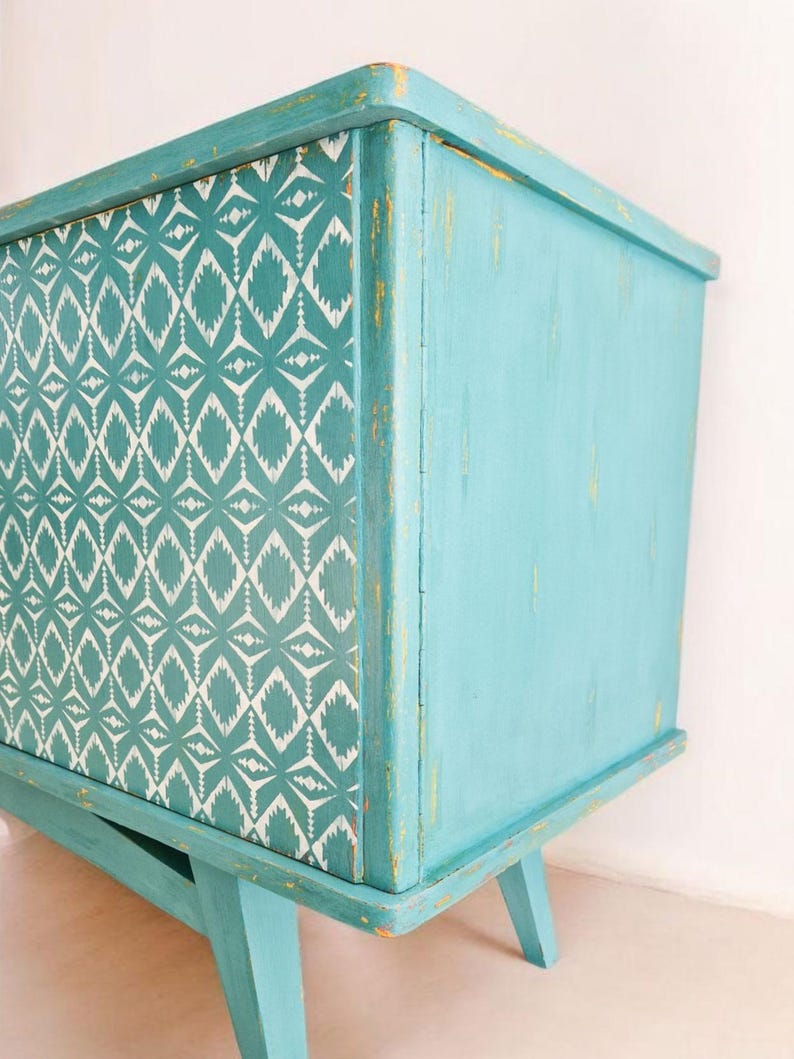 May include: A teal blue cabinet with a geometric pattern on the front. The cabinet has a distressed finish and sits on four legs.