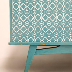 May include: A teal blue vintage cabinet with a geometric white pattern on the front. The cabinet has four legs that are angled outward.