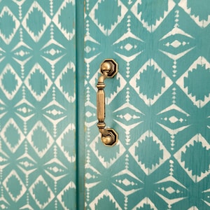 May include: A close-up of a teal cabinet door with a white geometric pattern. The cabinet door has a gold metal handle.