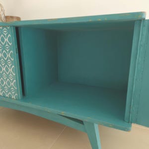 May include: Teal blue cabinet with a single door and a patterned panel on the other side. The cabinet has a simple design with angled legs.