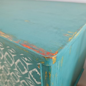 May include: A turquoise blue dresser with distressed paint and a white and turquoise geometric pattern on the front. The dresser has a yellow and orange accent color on the top edge.