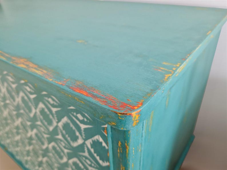 May include: A turquoise blue dresser with distressed paint and a white and turquoise geometric pattern on the front. The dresser has a yellow and orange accent color on the top edge.