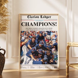 Puede incluir: Una impresión enmarcada de periódico con el titular "CHAMPIONS!" y el texto "OLE MISS FINISHES AMAZING RUN FROM LAST TEAM PICKED TO FIRST CWS TITLE." La imagen muestra a un equipo celebrando una victoria. El periódico se titula "Clarion Ledger."