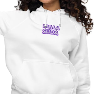 Puede incluir: Una sudadera blanca con capucha y mangas largas, que incluye un bolsillo tipo canguro frontal y cordones. En el pecho presenta el texto ME LA SUDA en letras moradas estilo burbuja con un borde de color morado oscuro.