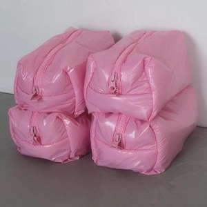 May include: A stack of four rectangular, light pink, glossy, padded pouches. Each pouch features a matching pink zip closure running along the top centre, with a single zip pull tab visible on the front end of each individual bag.