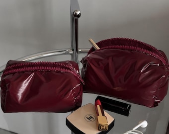 May include: Two glossy, dark burgundy zippered pouches are displayed alongside a square beige compact with a black Chanel logo, a gold-cased red lipstick, a black lipstick tube, and a pair of white wired earbuds on a glass surface.