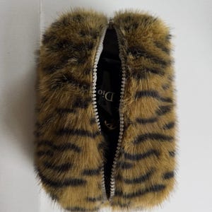 May include: A rectangular pouch covered in faux fur with a brown and black tiger stripe pattern. The pouch features a central silver-toned zip that is partially open, revealing a black interior lining with the visible white text Dior.