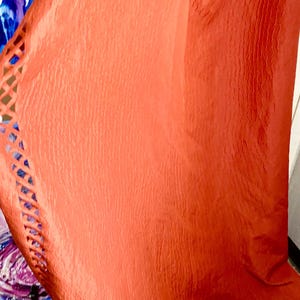 May include: A flowing, coral-colored fabric with a textured surface. The fabric has a decorative, openwork design along one edge, and a scalloped hemline. The material appears to be lightweight and drapes gracefully.