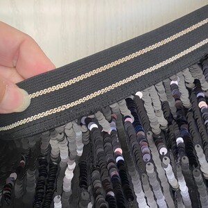 Op de afbeelding: Close-up van zwarte paillettenstof. De pailletten zijn rond en reflecteren licht, waardoor een glinsterend effect ontstaat. De tailleband is zwart met twee dunne, goudkleurige strepen. De stof wordt door een hand vastgehouden.