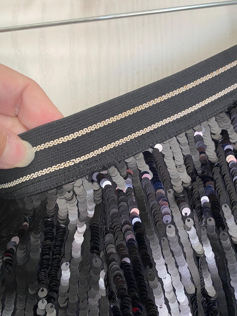 Op de afbeelding: Close-up van zwarte paillettenstof. De pailletten zijn rond en reflecteren licht, waardoor een glinsterend effect ontstaat. De tailleband is zwart met twee dunne, goudkleurige strepen. De stof wordt door een hand vastgehouden.