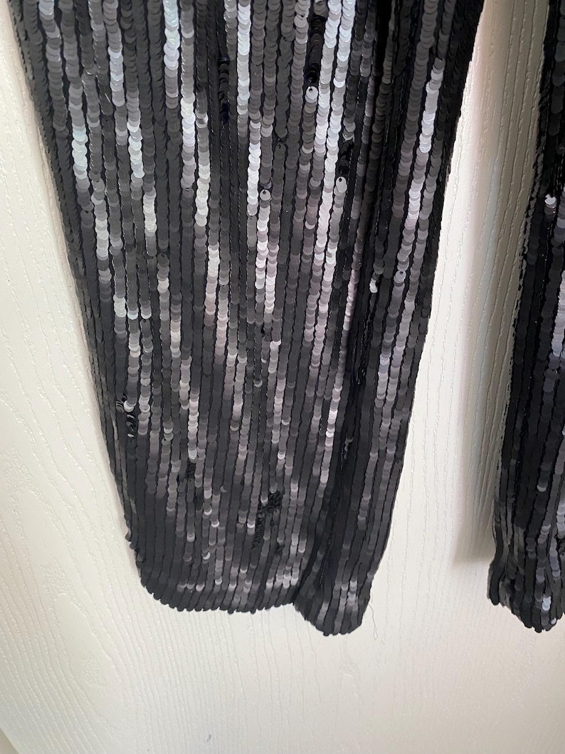 Op de afbeelding: Close-up van een zwarte paillettenbroek. De pailletten zijn verticaal uitgelijnd, waardoor een glinsterend effect ontstaat. De broek is gemaakt van een zachte, soepele stof, perfect voor een avondje uit of een speciale gelegenheid. De broek is effen donker van kleur.