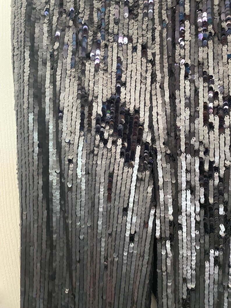 Op de afbeelding: Close-up van een kledingstuk bedekt met kleine, reflecterende, zilveren en grijze pailletten. De pailletten zijn gerangschikt in verticale rijen, waardoor een glinsterend, getextureerd oppervlak ontstaat. Het algehele effect is een glamoureus, opvallend ontwerp.