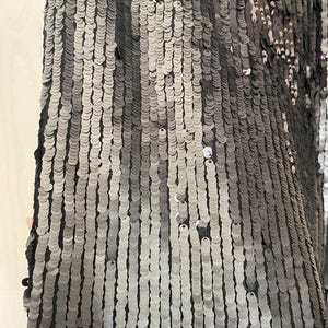 Op de afbeelding: Close-up van een stof bedekt met kleine, ronde, zilveren en grijze pailletten. De pailletten zijn gerangschikt in verticale rijen, waardoor een glinsterend effect ontstaat. De stof lijkt donkergrijs of zwart van kleur.