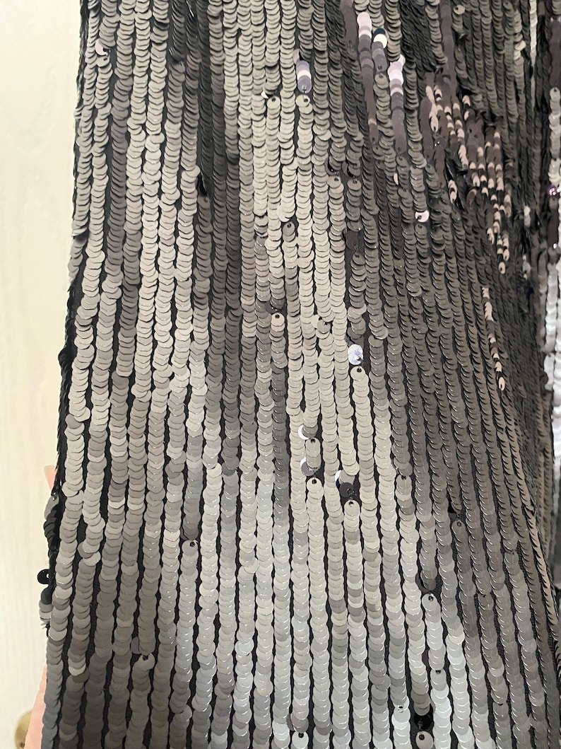 Op de afbeelding: Close-up van een stof bedekt met kleine, ronde, zilveren en grijze pailletten. De pailletten zijn gerangschikt in verticale rijen, waardoor een glinsterend effect ontstaat. De stof lijkt donkergrijs of zwart van kleur.