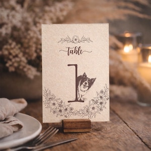 May include: A rustic table number card with the word "Table" in script and the number "1". The card features a floral design and a black and white dog illustration. It is displayed in a wooden holder.