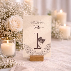 May include: A table number card with a floral design and the word "Table" in script. The number "1" features a dog peeking out. The card is held by a small wooden block, perfect for weddings or events.