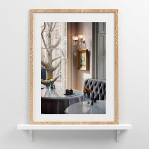 Op de afbeelding: Een ingelijste prent van een restaurantinterieur. De afbeelding toont een ronde tafel met een grijs tafelkleed, een gecapitonneerde grijze bank en een raam met uitzicht op een boom. Een klein schilderij hangt aan de muur en een wandlamp zorgt voor zachte verlichting.