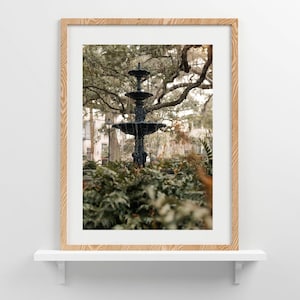 Op de afbeelding: Een ingelijste print met een gelaagde fontein in een park. De donkere fontein staat tegen weelderig groen en grote bomen. De print zit in een licht houten frame, tentoongesteld op een witte plank tegen een witte muur.