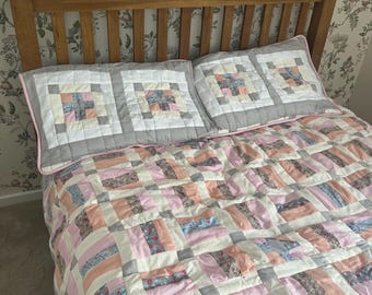 May include: A handmade quilt and two matching pillow shams on a bed. The quilt features a patchwork design with pink, peach, blue, and floral fabric blocks. The pillow shams have a square pattern with gray borders. The bed has a wooden headboard.