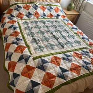 May include: A quilted bedspread featuring a geometric pattern of orange, navy blue, and cream fabric squares. The centre panel is framed by a solid olive green border, with additional orange and navy blue squares forming the outer edges.