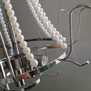 May include: A silver-toned metal chandelier with multiple curved arms and hooks. Strands of white faux pearls are draped across the fixture. The design is modern and elegant, suitable for home decor.