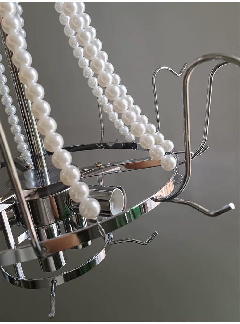 May include: A silver-toned metal chandelier with multiple curved arms and hooks. Strands of white faux pearls are draped across the fixture. The design is modern and elegant, suitable for home decor.