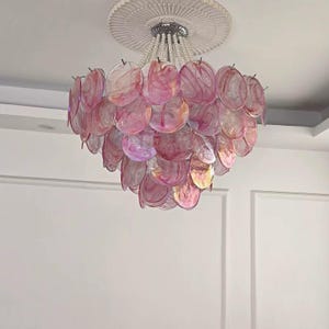 May include: A decorative chandelier featuring a cascade of pink, translucent, shell-shaped elements. The light fixture is suspended from a silver-toned canopy with beaded strands, and is mounted on a white ceiling.
