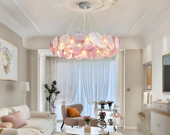 May include: A pink and white glass chandelier with a circular design hangs from the ceiling in a living room. The chandelier is made up of many individual glass pieces that are arranged in a circular pattern. The chandelier is lit and casts a warm glow on the room.