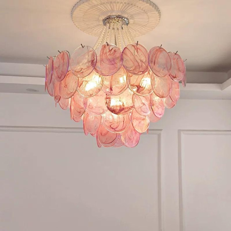 May include: A pink and clear chandelier with a layered design. The light fixture is suspended from the ceiling and features multiple circular, petal-like elements. The chandelier has a silver-colored base and is illuminated by several lights.