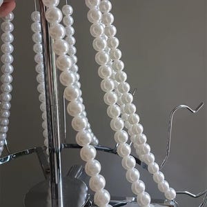 May include: Close-up of a chandelier with multiple strands of white faux pearls. The chandelier has a silver metal frame with curved arms and a central support. The pearls are strung together, creating a cascading effect.