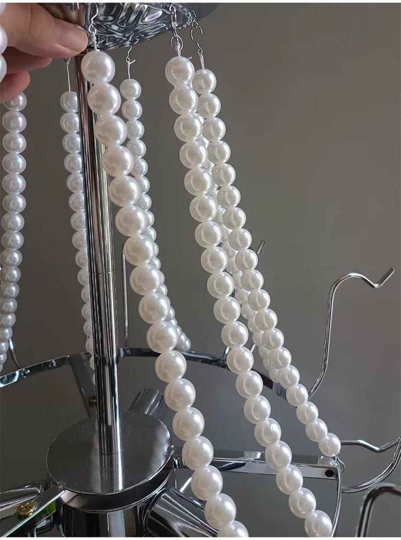 May include: Close-up of a chandelier with multiple strands of white faux pearls. The chandelier has a silver metal frame with curved arms and a central support. The pearls are strung together, creating a cascading effect.