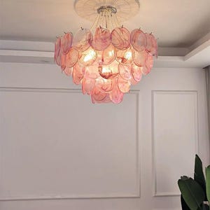May include: A decorative chandelier with a layered design of pink, translucent, petal-shaped elements. The light fixture hangs from a white ceiling with a circular, textured trim. The chandelier's lights are illuminated.