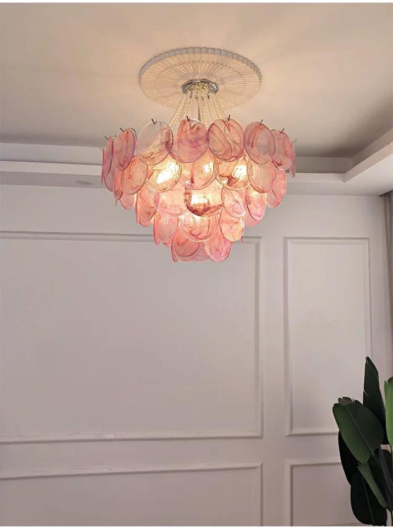 May include: A decorative chandelier with a layered design of pink, translucent, petal-shaped elements. The light fixture hangs from a white ceiling with a circular, textured trim. The chandelier's lights are illuminated.